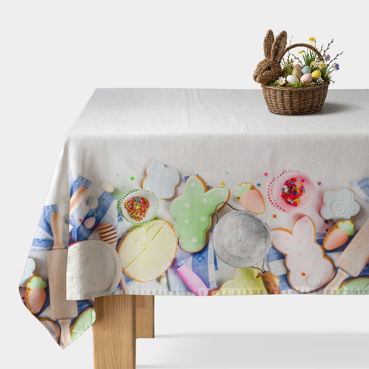 Obrus rozłożony na stole z kolorowym brzegiem przedstawiającym pisanki i lukrowane ciasteczka oraz wiklinowy koszyk z dekoracyjnymi jajkami w rogu