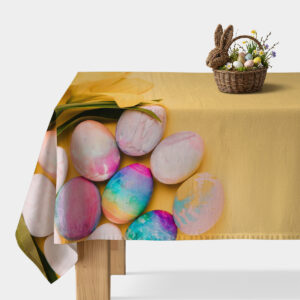 Obrus z żółtym tłem i pastelowymi pisankami ułożonymi przy krawędzi stołu, na blacie koszyk z dekoracyjnymi jajkami