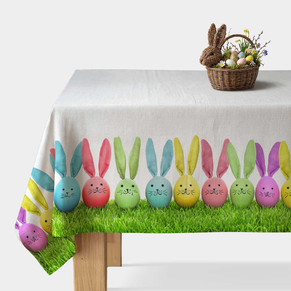 Obrus na białym stole z nadrukiem pastelowych pisanek z uszami zajączków stojących na zielonej trawie