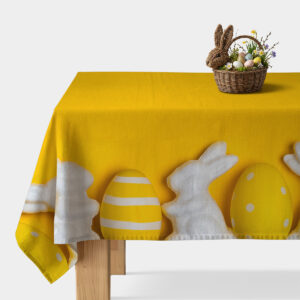 Główne ujęcie: żółty obrus z białymi sylwetkami zajączków i ozdobnymi pisankami, stół z koszykiem dekoracji