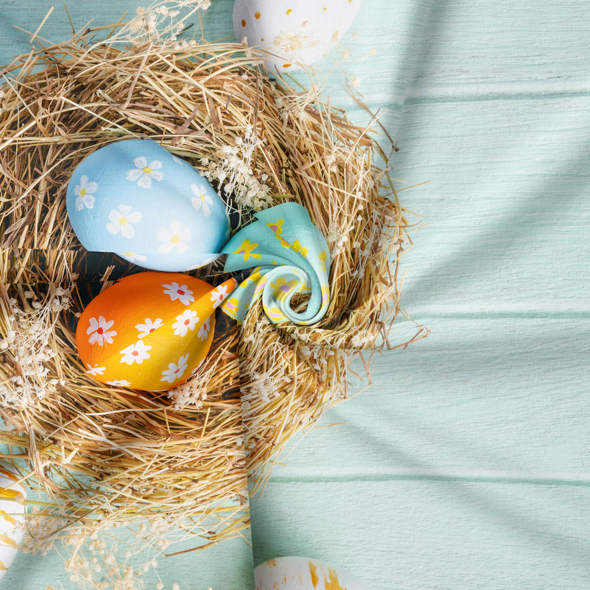 zbliżenie na gniazdo ze słomą i pastelowymi pisankami na tle miętowego materiału