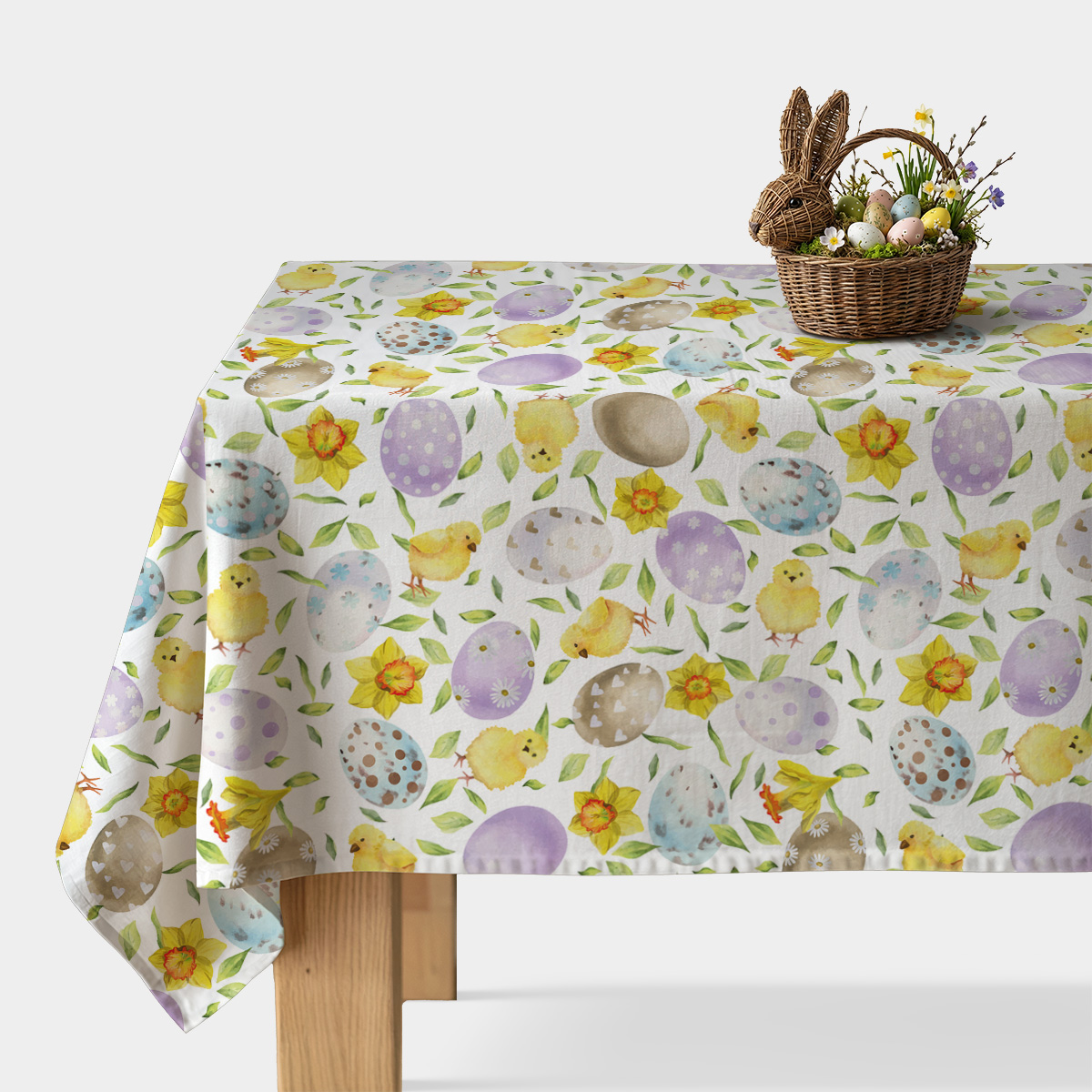 stół nakryty obrusem w akwarelowe pisanki i kurczaczki z wiklinowym koszykiem na jajka