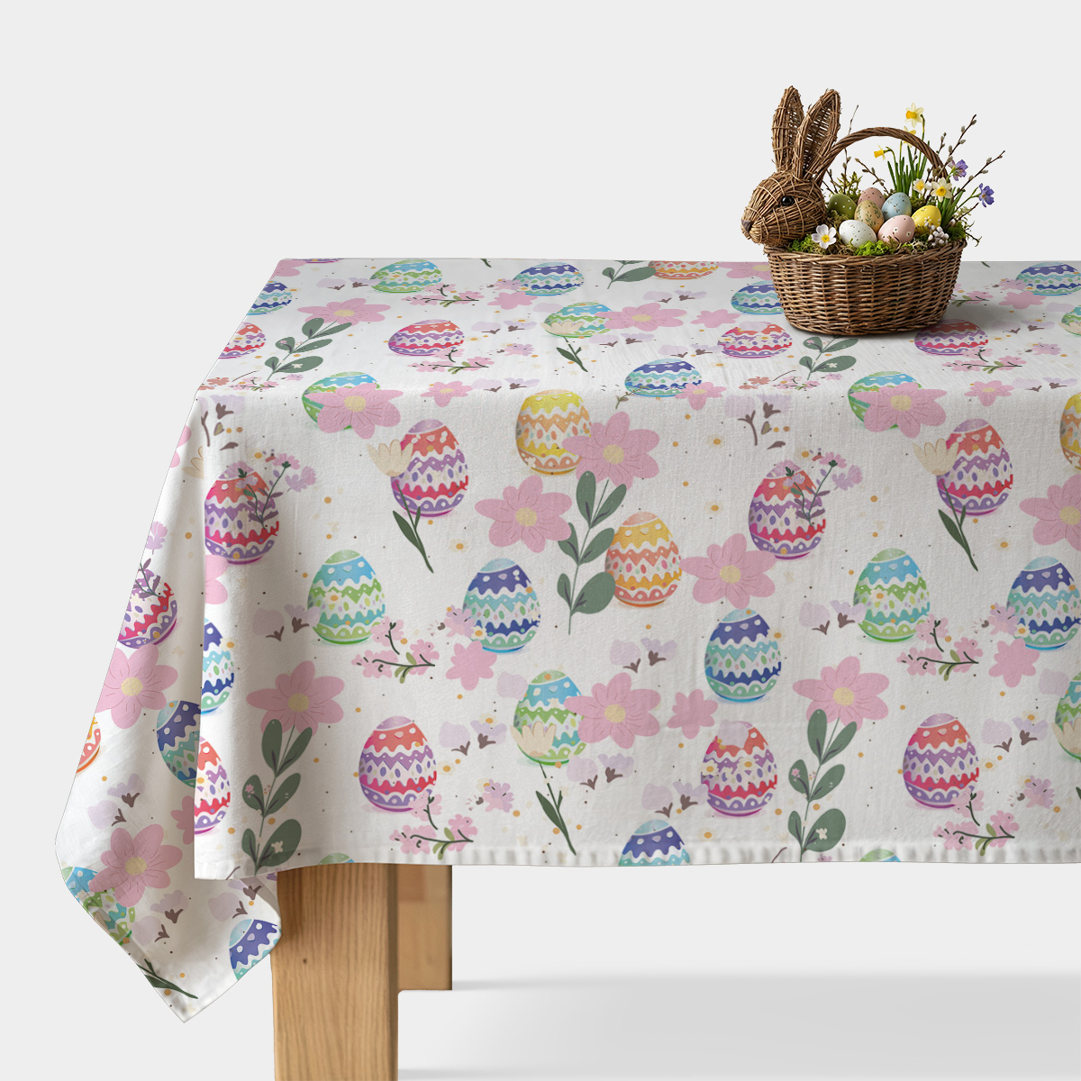 Stół nakryty obrusem z pastelowymi pisankami i różowymi kwiatami oraz koszyk wielkanocny z dekoracyjnymi jajkami