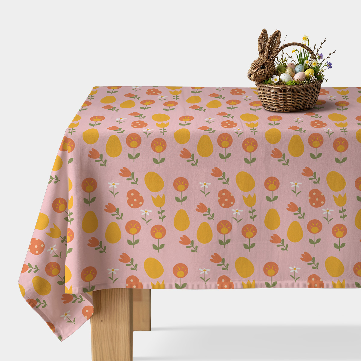 stół z różowym obrusem w motyw pisanek i kwiatów z wiklinowym koszykiem wielkanocnym