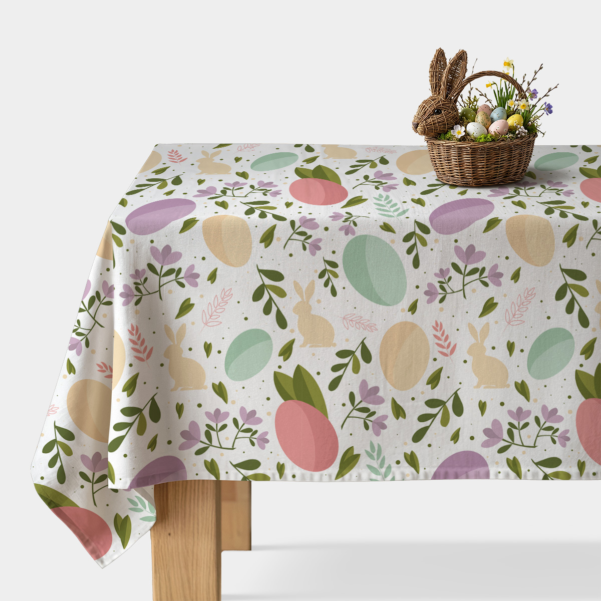 Stół nakryty obrusem z pastelowymi pisankami i zajączkami oraz wiklinowy koszyk z dekoracyjnymi jajkami