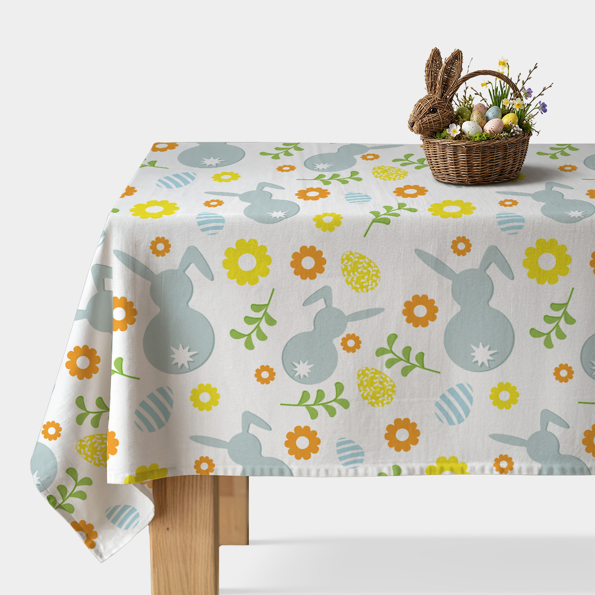 Stół przykryty obrusem z wzorem zajączków i pisanek, na rogu koszyk z wielkanocnymi jajkami