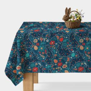 Stół nakryty obrusie z motywem zajączków i kwiatów, na blacie wiklinowy koszyk z pisankami i dekoracyjnym króliczkiem