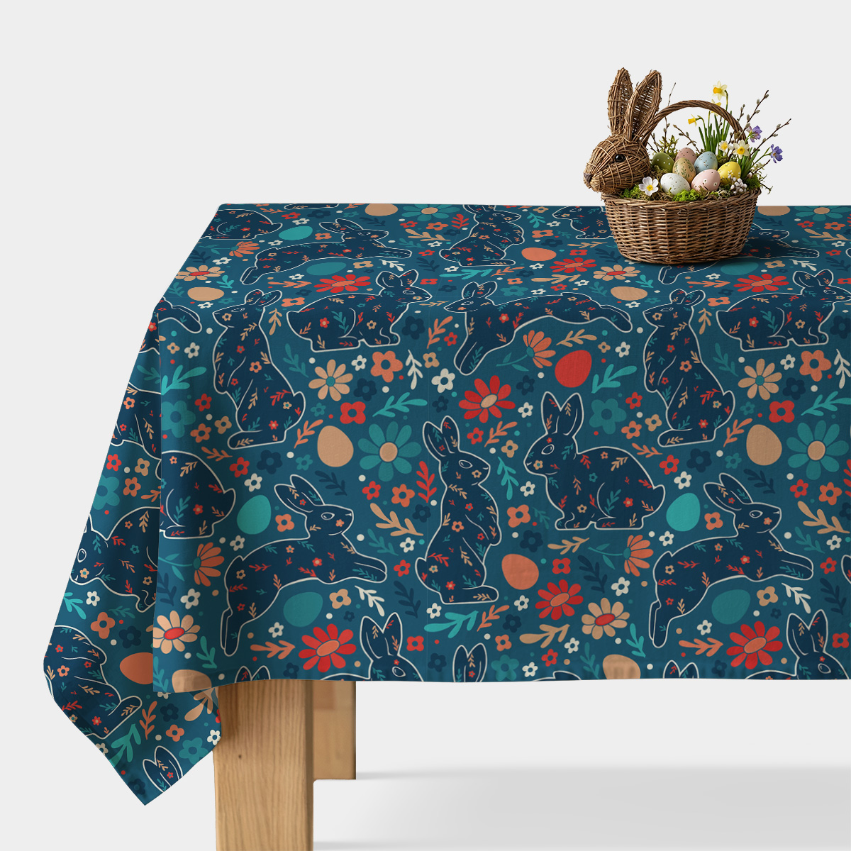 Stół nakryty obrusie z motywem zajączków i kwiatów, na blacie wiklinowy koszyk z pisankami i dekoracyjnym króliczkiem