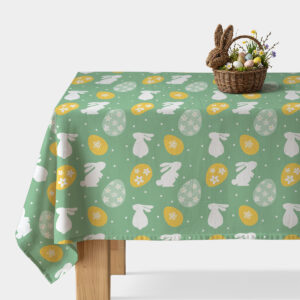 Obrus w miętowym odcieniu z białymi zajączkami i żółtymi pisankami ułożony na stole z dekoracyjnym koszykiem