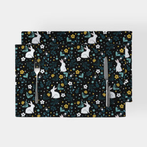 Dwie prostokątne podkładki na czarnym tle z wzorem białych zajączków i drobnymi kwiatami, ustawione obok sztućców