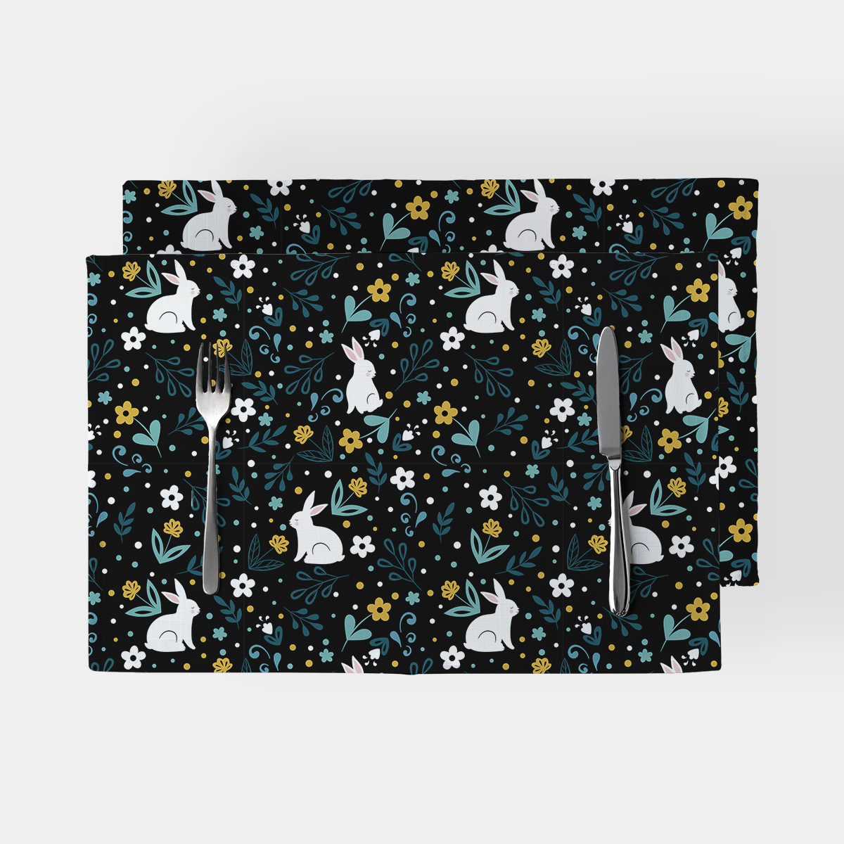 Dwie prostokątne podkładki na czarnym tle z wzorem białych zajączków i drobnymi kwiatami, ustawione obok sztućców