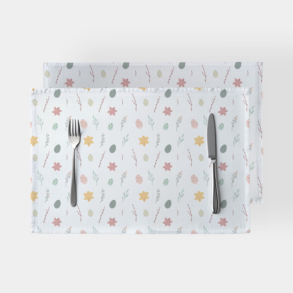 Dwie podkładki na stół z pastelowym wzorem pisanek i wiosennych gałązek ułożone jedna na drugiej ze sztućcami