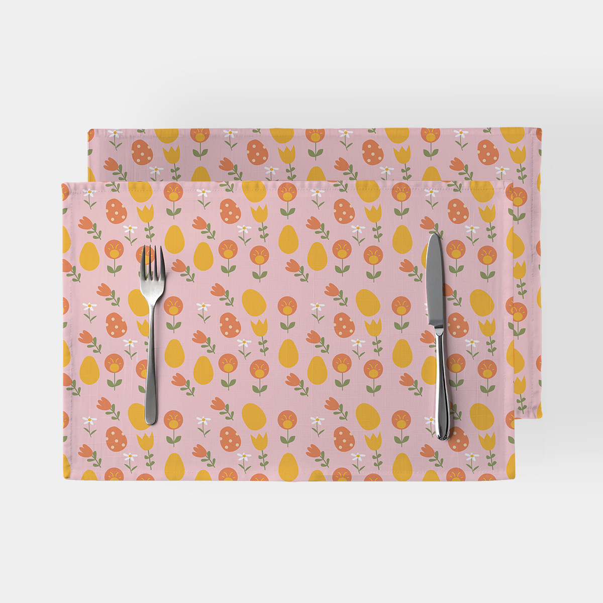 Dwie podkładki na stół z różowym tłem ozdobione żółtymi pisankami i pomarańczowymi kwiatami, ujęcie z widocznym sztućcem