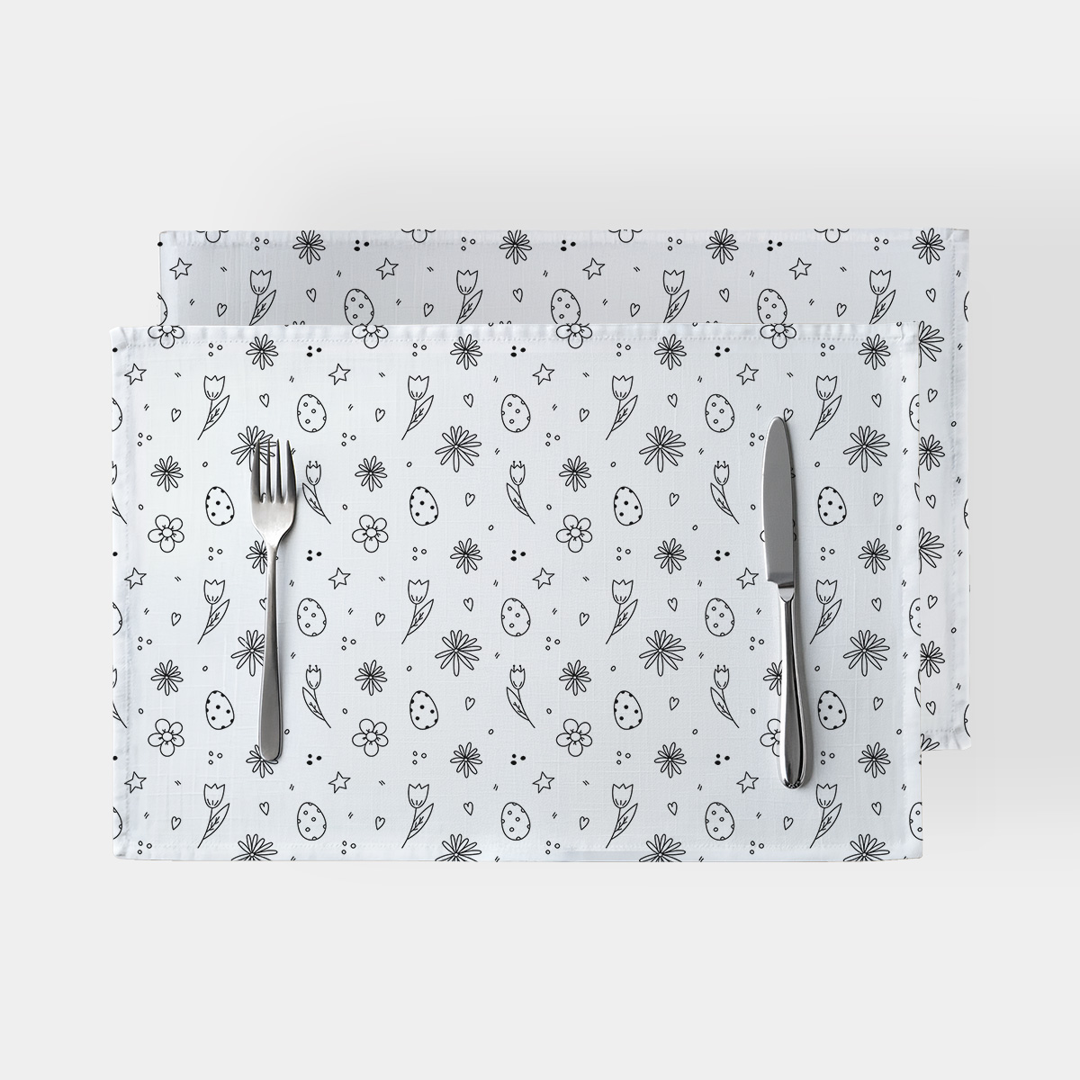Para białych podkładek z czarno-białym wzorem pisanek i kwiatów na jasnym tle z widocznymi sztućcami