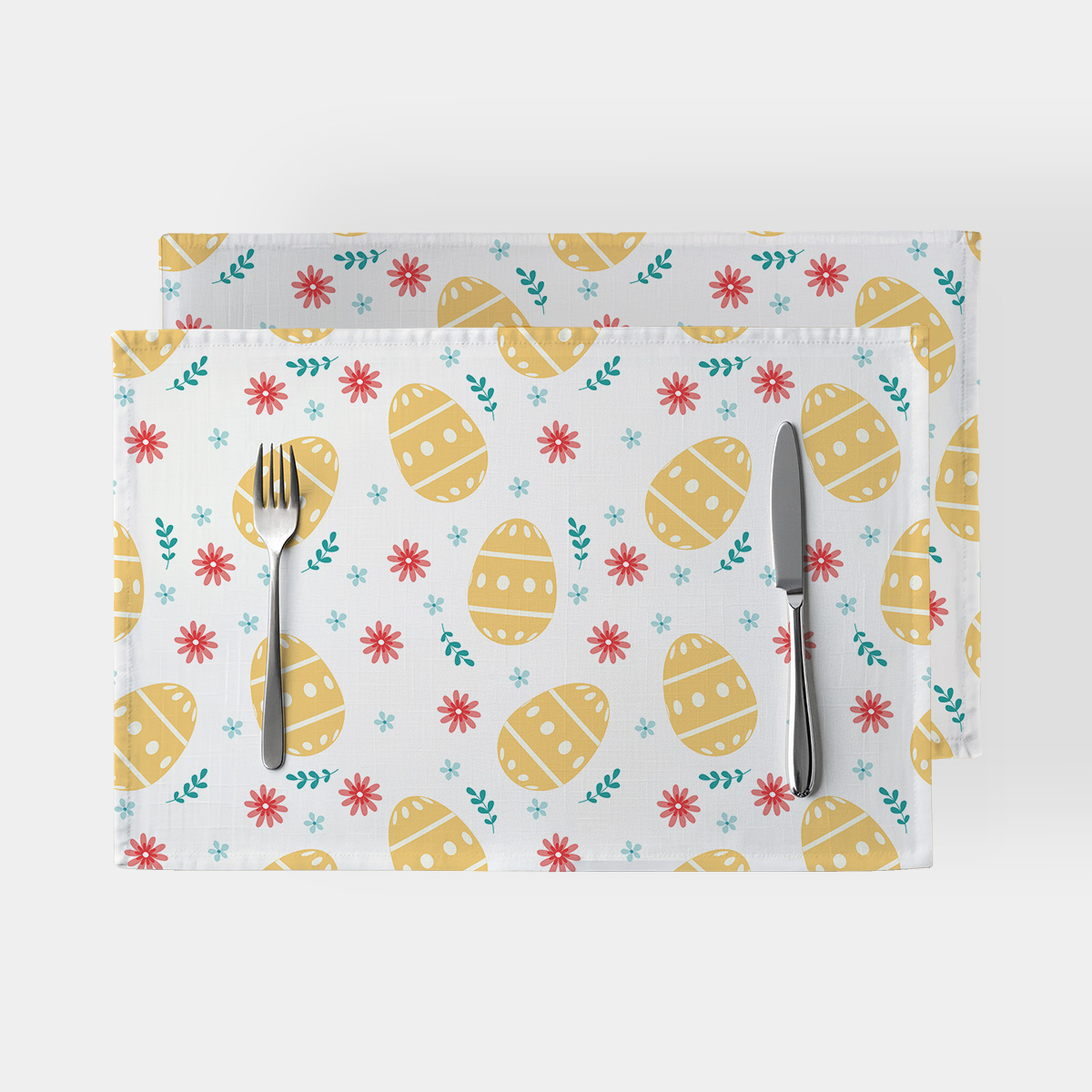 Dwie podkładki z motywem żółtych pisanek i drobnych czerwonych oraz błękitnych kwiatów ułożone na jasnym tle ze sztućcami