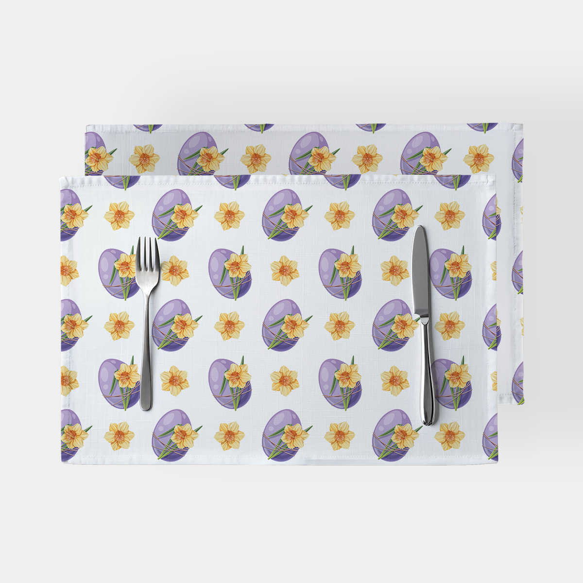 Główne ujęcie dwóch białych podkładek ze wzorem fioletowych pisanek i żółtych żonkili ułożonych z sztućcami