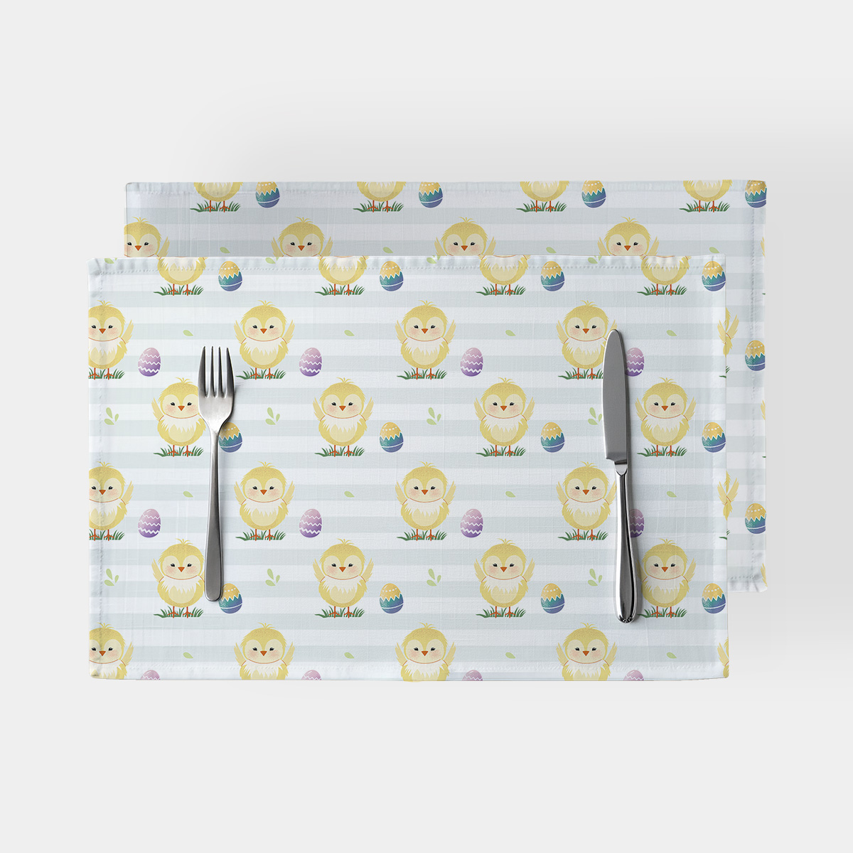 Widok z góry dwóch podkładek na stół z wzorem uśmiechniętych kurczaczków i pisanek na jasnym, paskowanym tle