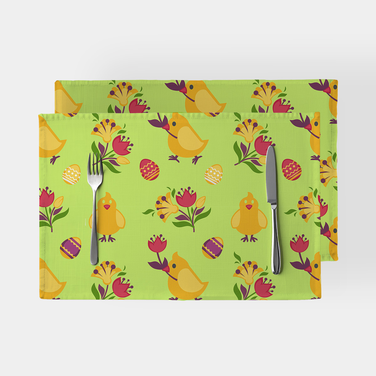 Dwie zielone podkładki na stół z motywem żółtych kurczaczków, pisanek i kwiatów ułożone z nożem i widelcem