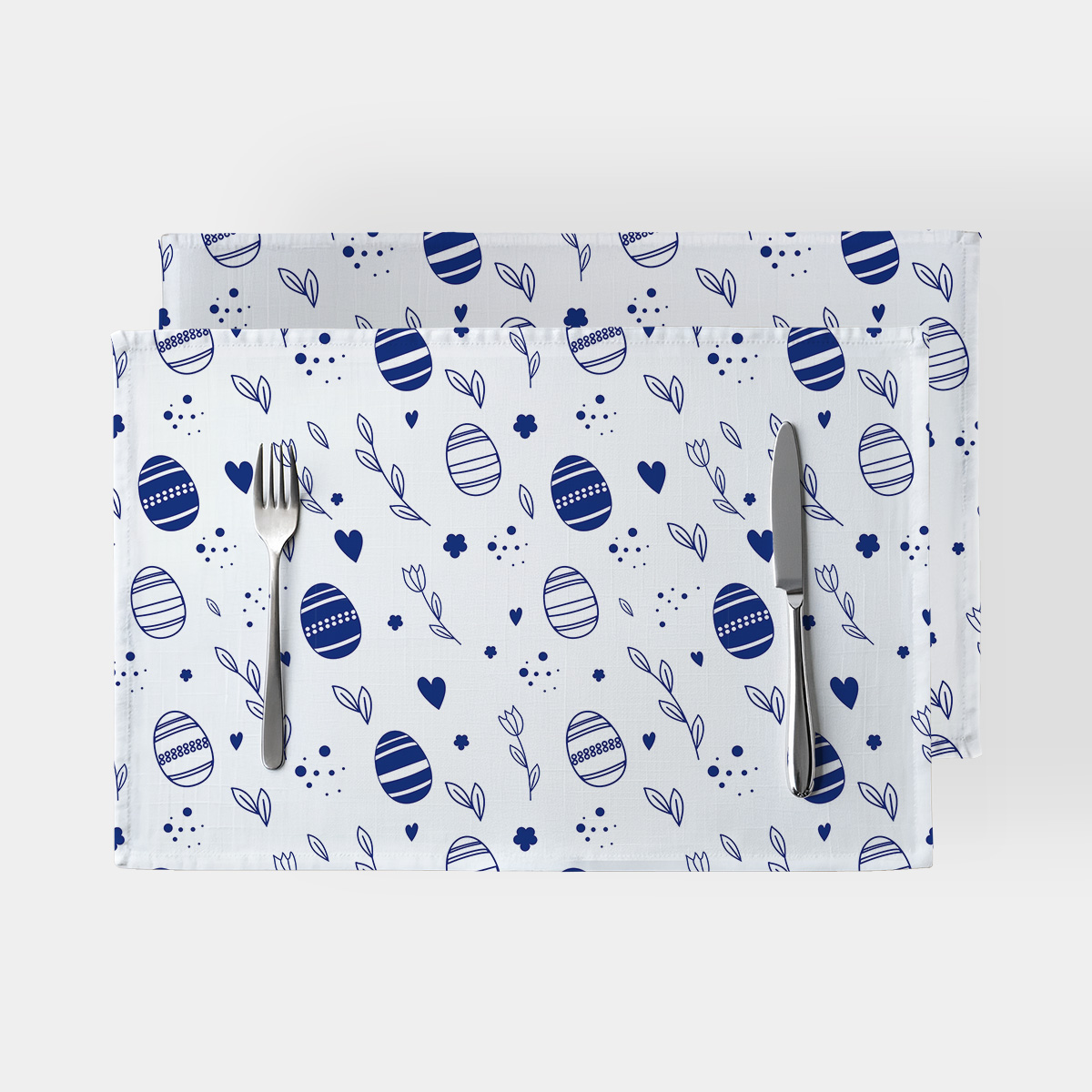 Dwie prostokątne podkładki na białym tle z granatowym motywem pisanek i gałązek, ułożone ze sztućcami