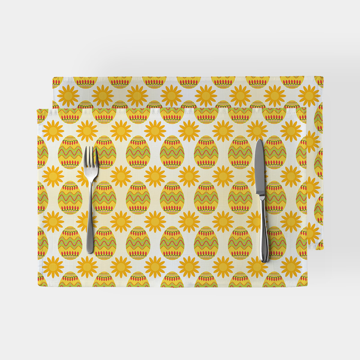 Dwie podkładki na stół z wzorem wielkanocnych pisanek i żółtych słoneczek na jasnym tle z widelcem i nożem ułożonymi na produkcie