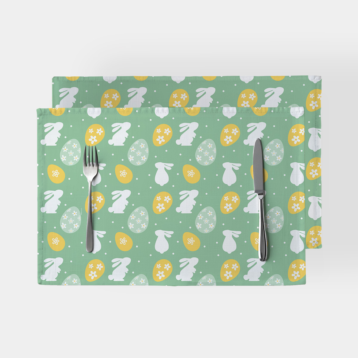 Para podkładek w zielonym kolorze z białymi zajączkami i żółtymi pisankami ułożona jedna na drugiej z widocznym nożem i widelcem