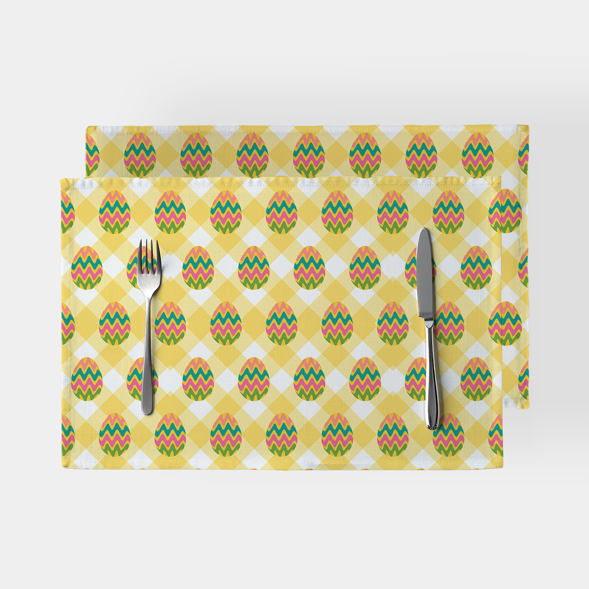 Dwie podkładki w żółtą kratę z kolorowymi pisankami ułożone na stole z nożem i widelcem, widok z góry
