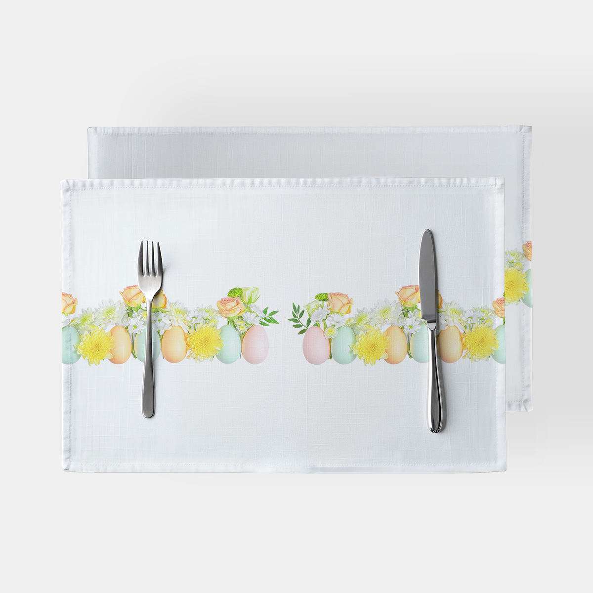 biała podkładka z poziomym paskiem pastelowych jajek i żółtych kwiatów, ujęcie z góry z widocznymi sztućcami