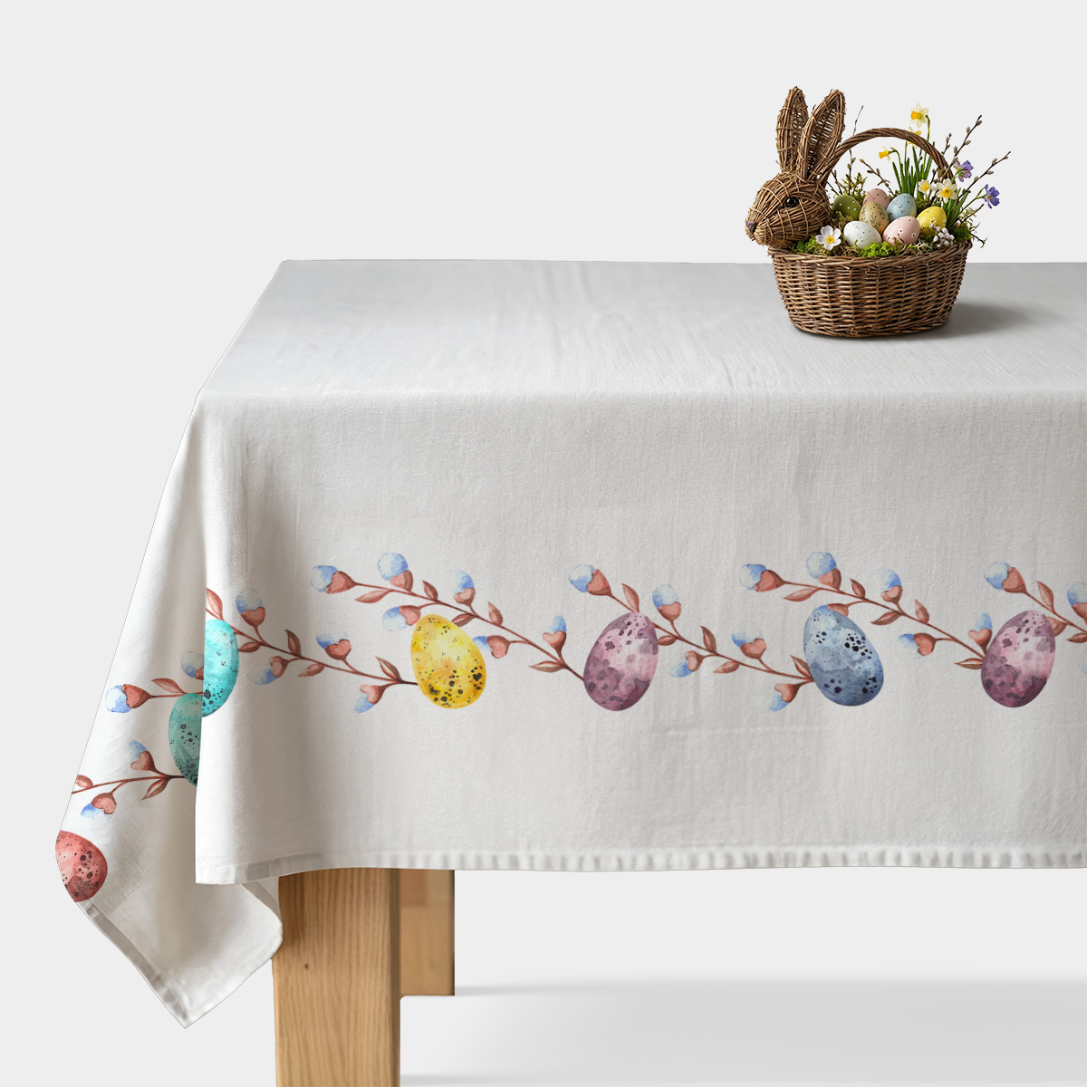 biały obrus z pasem akwarelowych pisanek i gałązek na stole z dekoracyjnym koszykiem