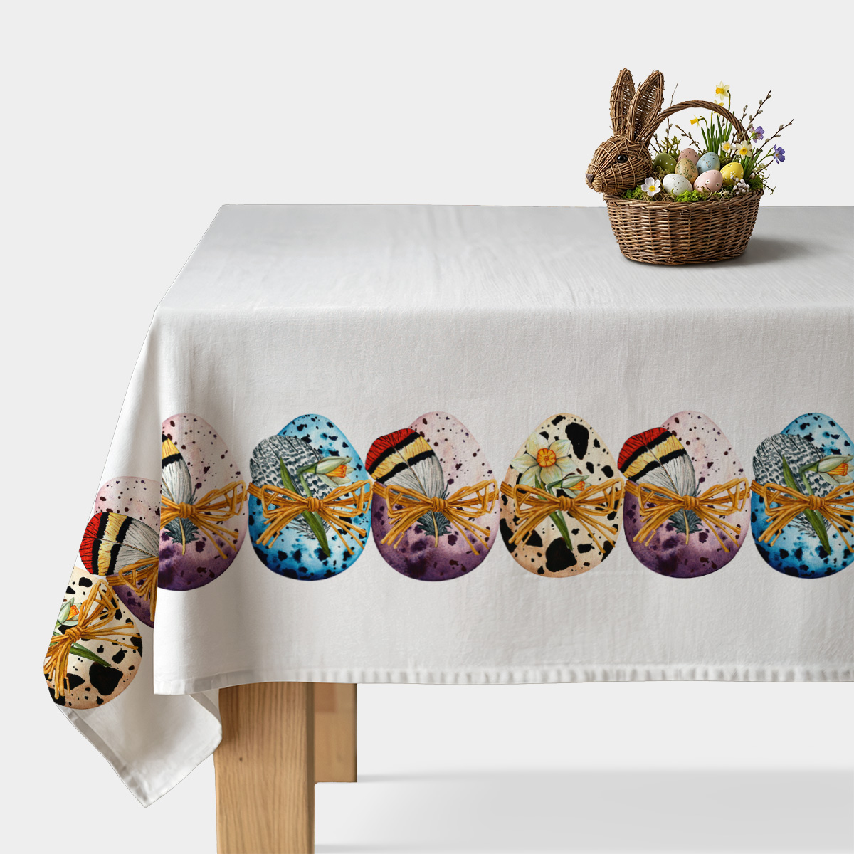 biały obrus z dekoracyjnym pasem malowanych pisanek i wiklinowy koszyk z jajkami ustawiony na stole