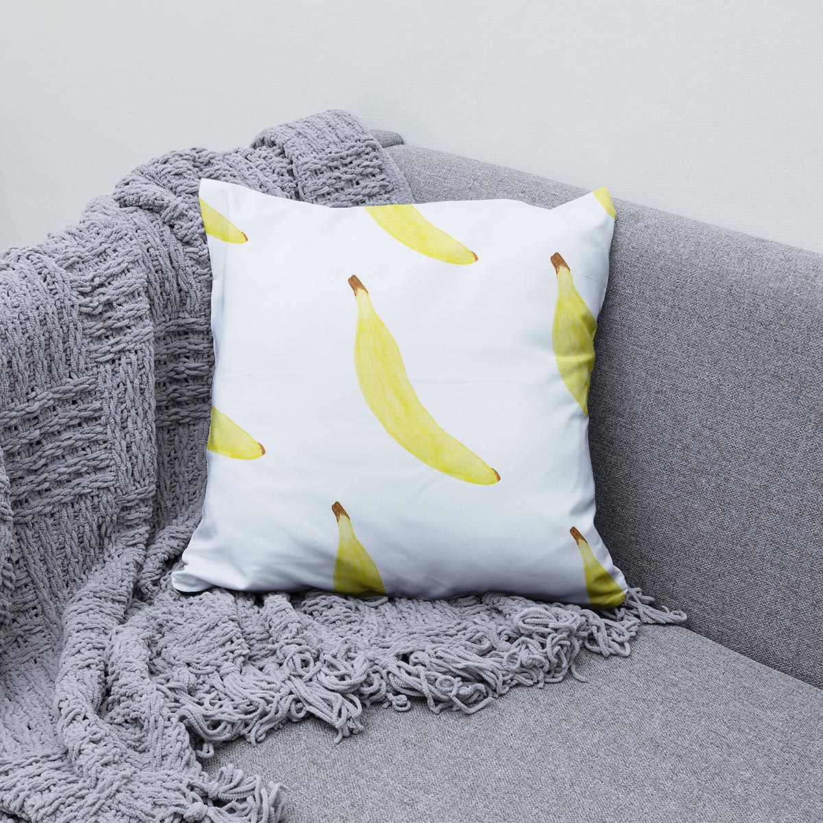 Poduszka z bananowym wzorem ułożona na szarej sofie i pledzie jako przytulna aranżacja