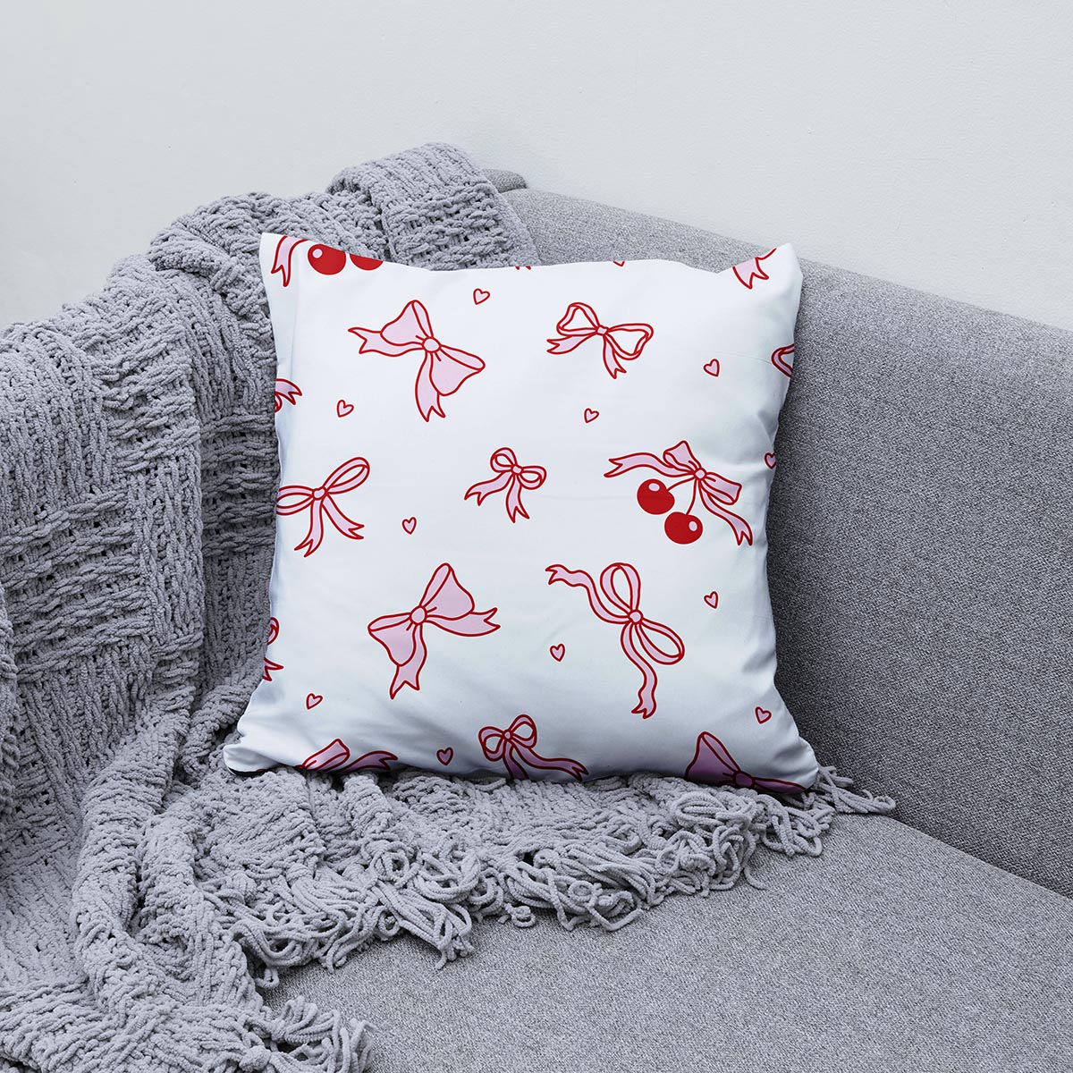 Poduszka dekoracyjna z motywem kokardek i wiśni ułożona na kanapie z pledem