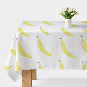 Obrus z akwarelowymi bananami rozłożony na stole, wazon z białymi kwiatami w tle