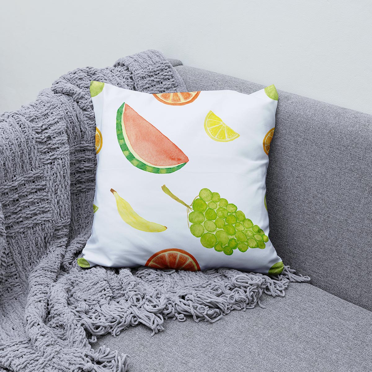 Poduszka ozdobna na kanapie obok szarego koca, motyw banana i plasterków cytrusów