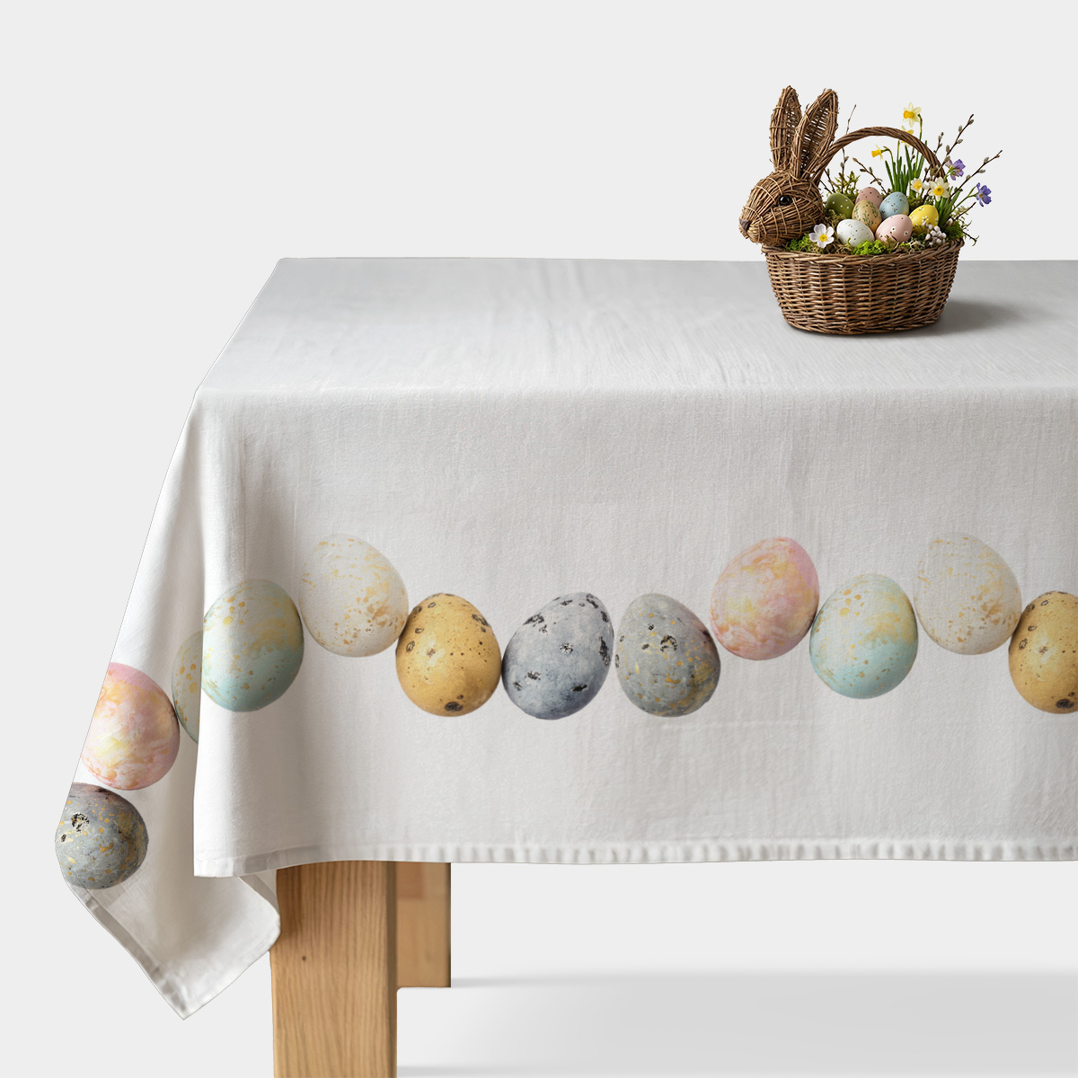 Biały obrus na stole z pasem pastelowych pisanek i złotymi plamkami, udekorowany koszykiem z jajkami