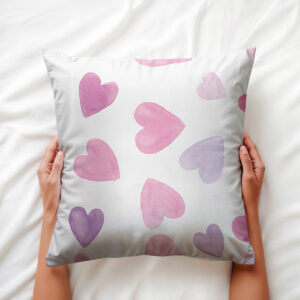 Poduszka ozdobna z akwarelowymi sercami trzymana w dłoniach na białej pościeli