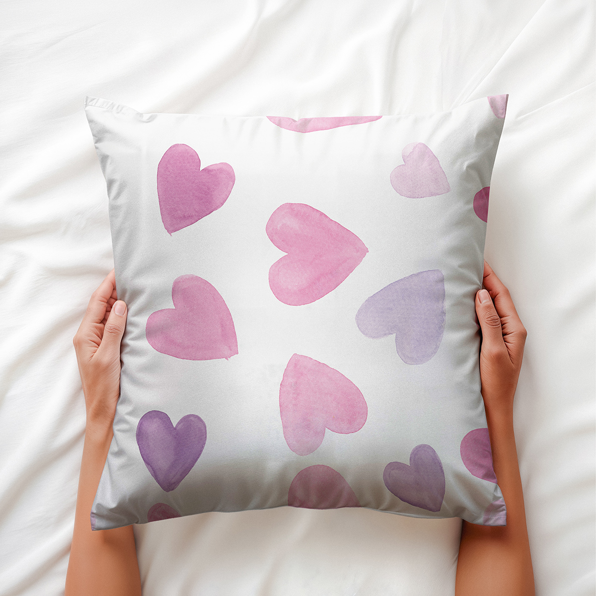Poduszka ozdobna z akwarelowymi sercami trzymana w dłoniach na białej pościeli