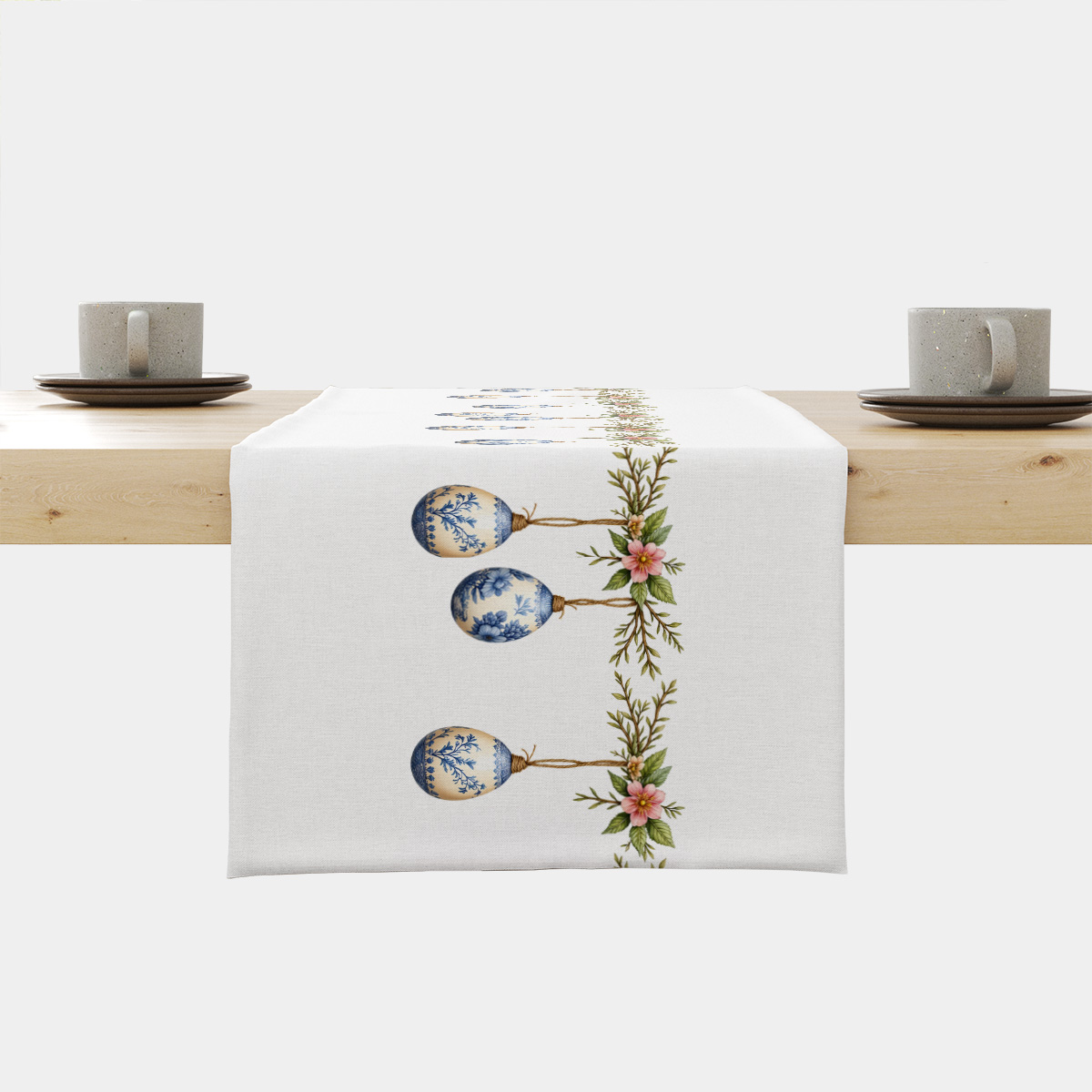 biały bieżnik z wiszącymi niebiesko-kremowymi porcelanowymi pisankami i gałązkami ułożony na drewnianym stole