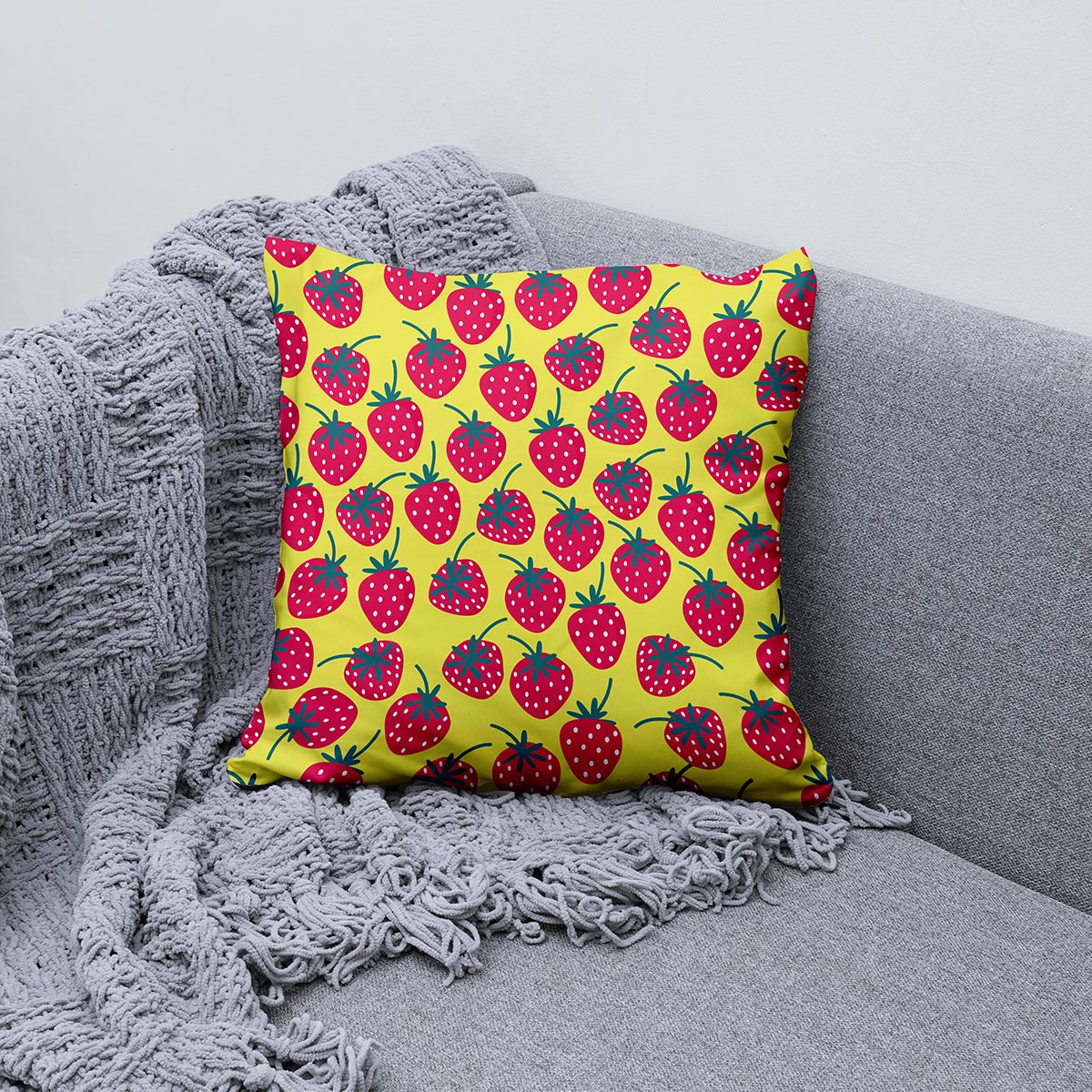 Poduszka z wzorem truskawek ułożona na szarej sofie obok pledu, detal wzoru