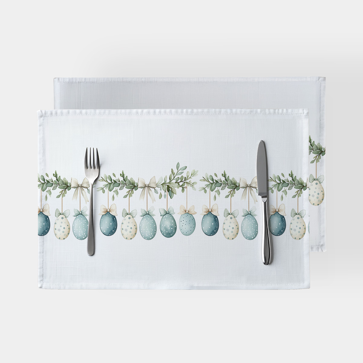 Podkładki ze wzorem wiszących pisanek i kokard na białym tle, ujęcie z widocznym sztućcem