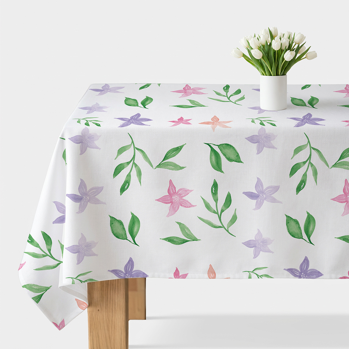 Obrus z akwarelowymi fioletowymi i różowymi kwiatami oraz zielonymi listkami na białym tle, główne ujęcie stołu z wazonem białych tulipanów