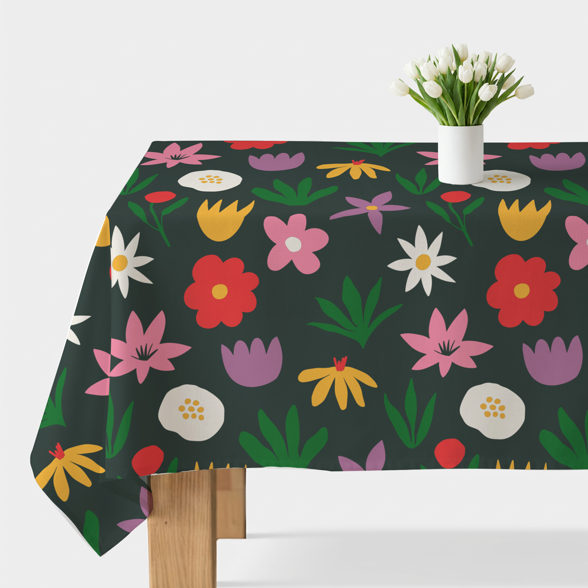 Stół przykryty obrusem z ciemnym tłem i wesołym wzorem kolorowych kwiatów, na środku biały wazon z tulipanami