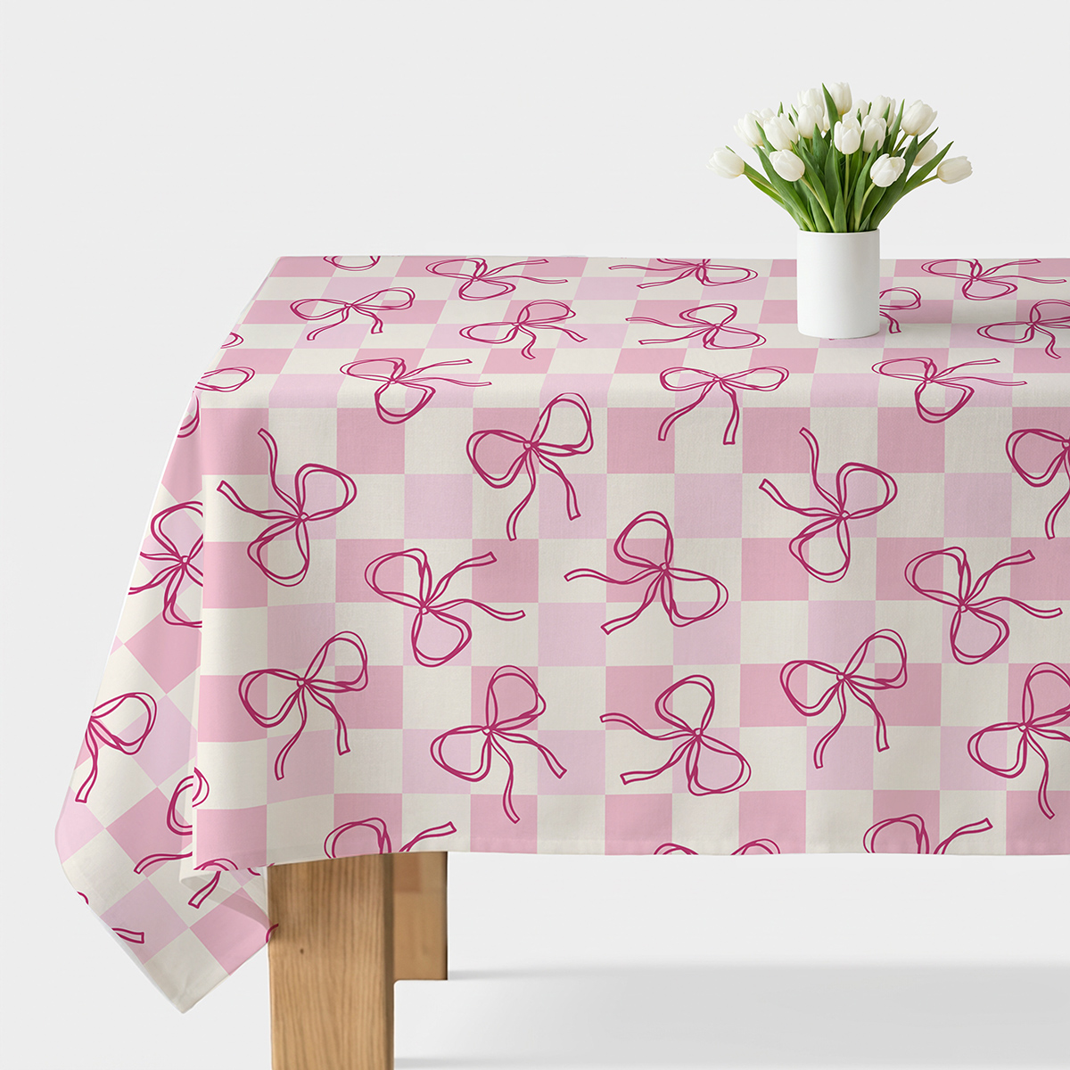 obrus z pudrowo-kremową kratką i malowanymi różowymi kokardkami rozłożony na stole z wazonem białych tulipanów
