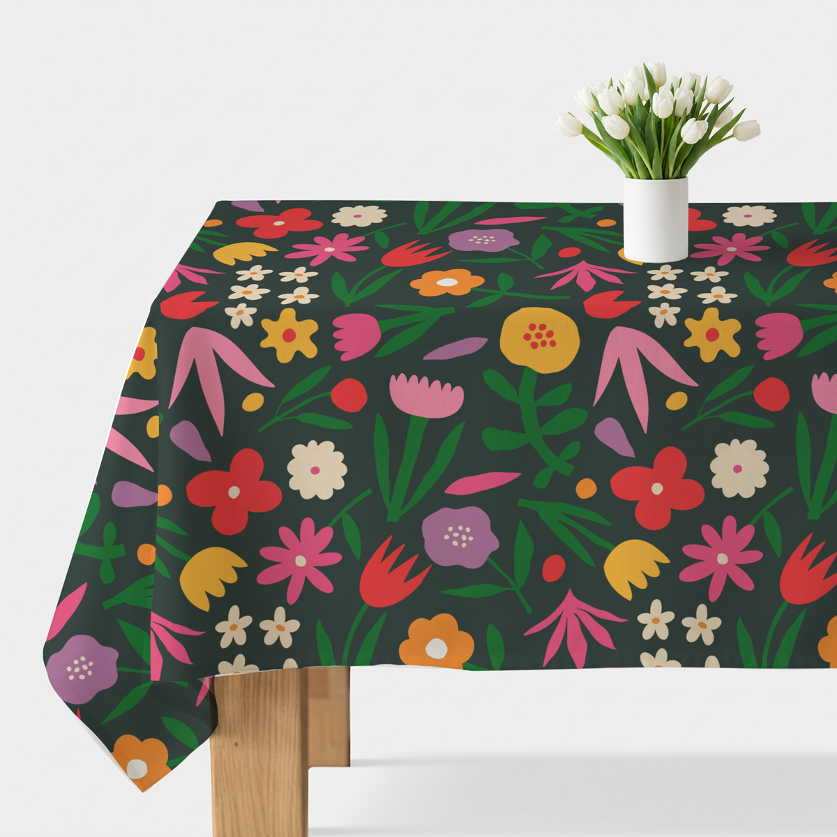 Obrus rozłożony na stole z kolorowym, stylizowanym wzorem kwiatowym na ciemnozielonym tle i wazonem z białymi tulipanami
