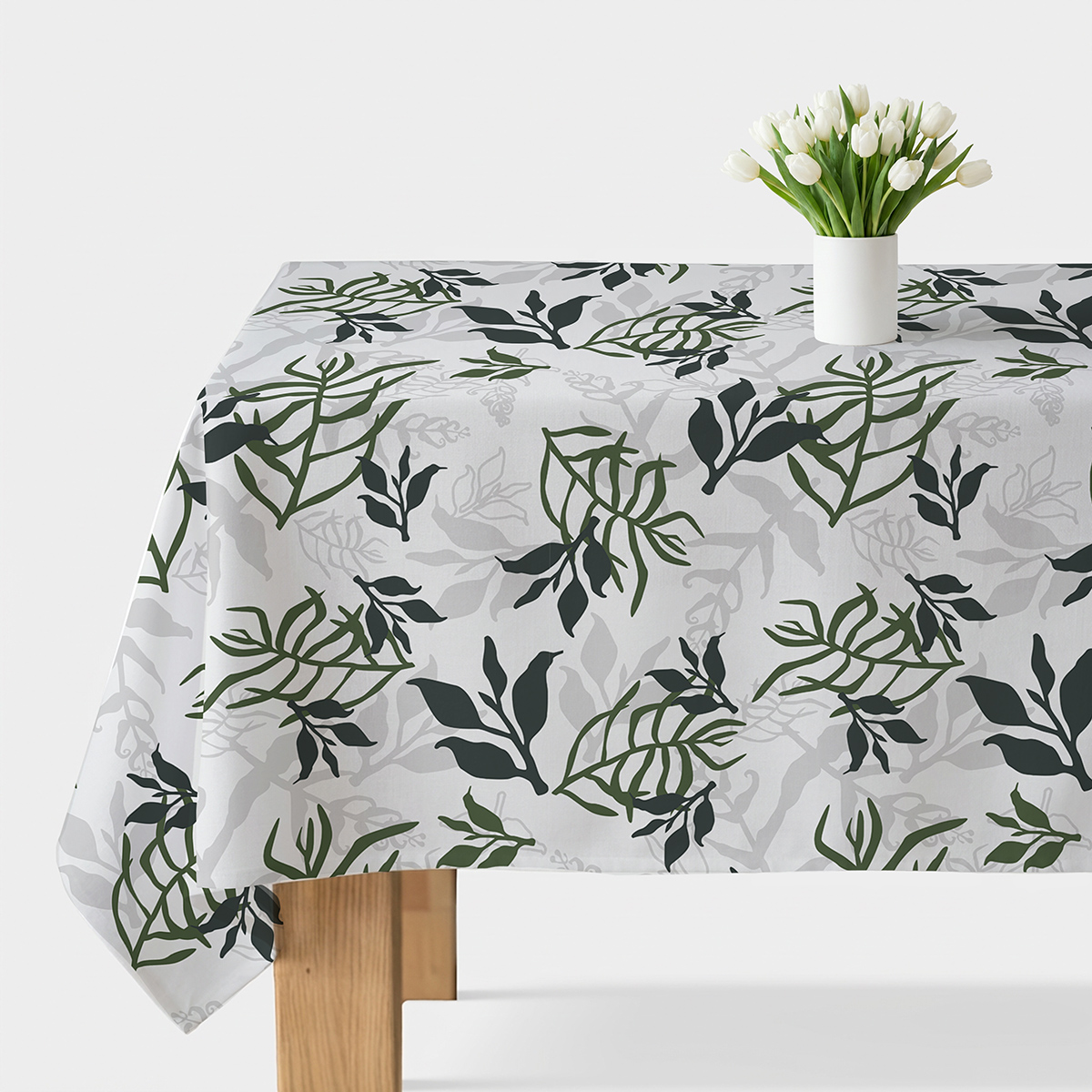 Stół nakryty obrusem z jasnym tłem i zielonym liściastym wzorem, na środku biały wazon z tulipanami