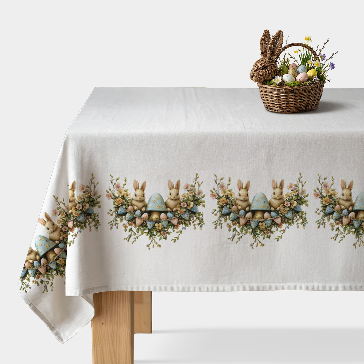 Obrus ze świątecznym motywem zajączków, pisanek i kwiatów na białym tle, widok stołu z koszykiem dekoracji