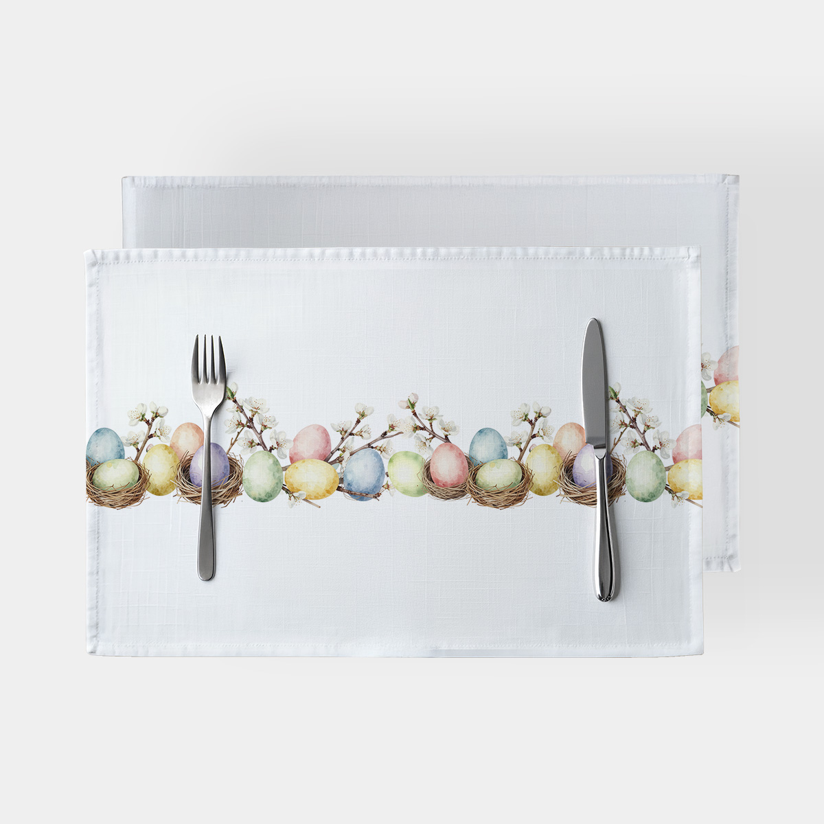 Podkładki obiadowe na białym tle z wzorem pastelowych pisanek i kwitnących gałązek, ustawione ze sztućcami