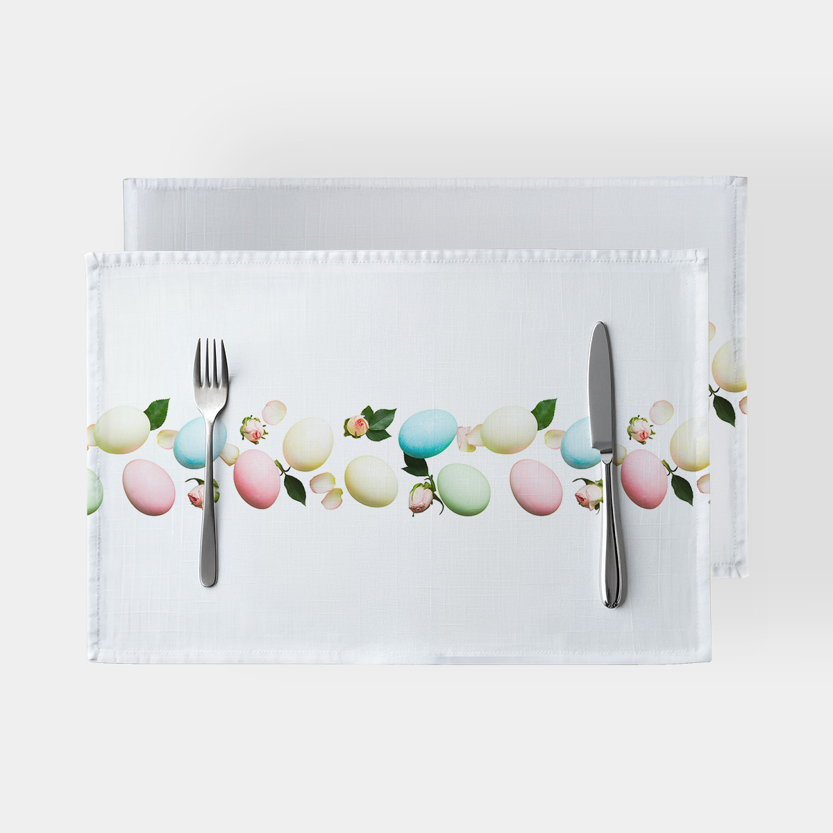 Widok z góry na dwie świąteczne podkładki z pasem pastelowych pisanek i pąków róż oraz sztućce