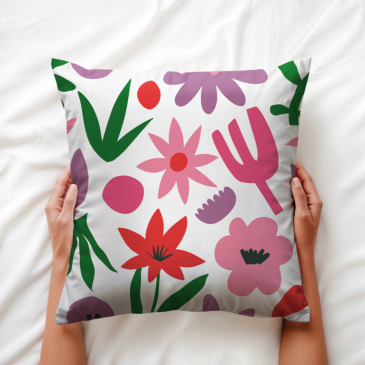Poduszka trzymana w dłoniach na białej pościeli, wzór z różowymi i czerwonymi kwiatami oraz zielonymi liśćmi