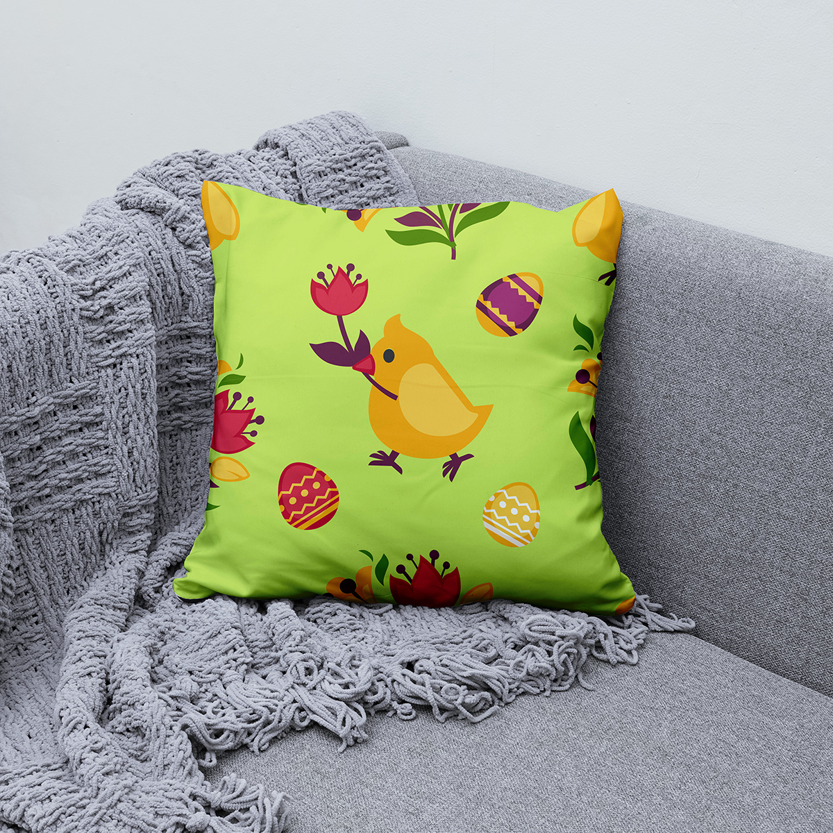 Poduszka ułożona na szarej sofie obok pledu, widoczny cały wzór z kurczakiem i pisankami