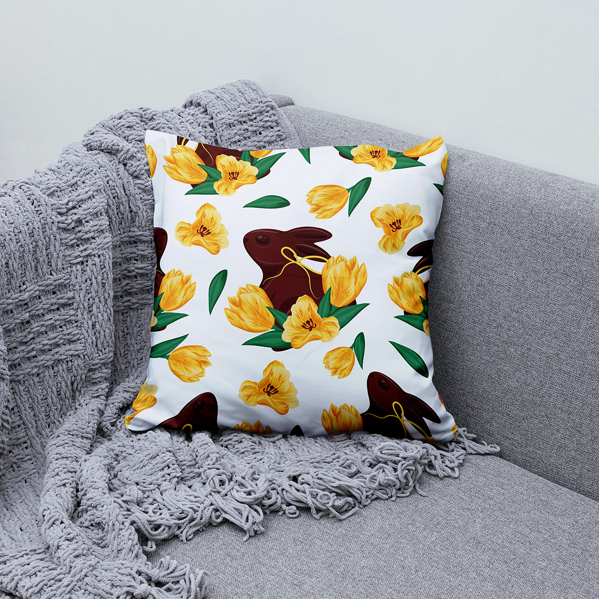 Poduszka z wzorem zająca i żółtych tulipanów ułożona na kanapie przy szarym kocu